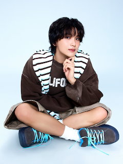 Young man with dark hair sitting cross-legged wearing brown jacket, striped shirt, and blue sneakers