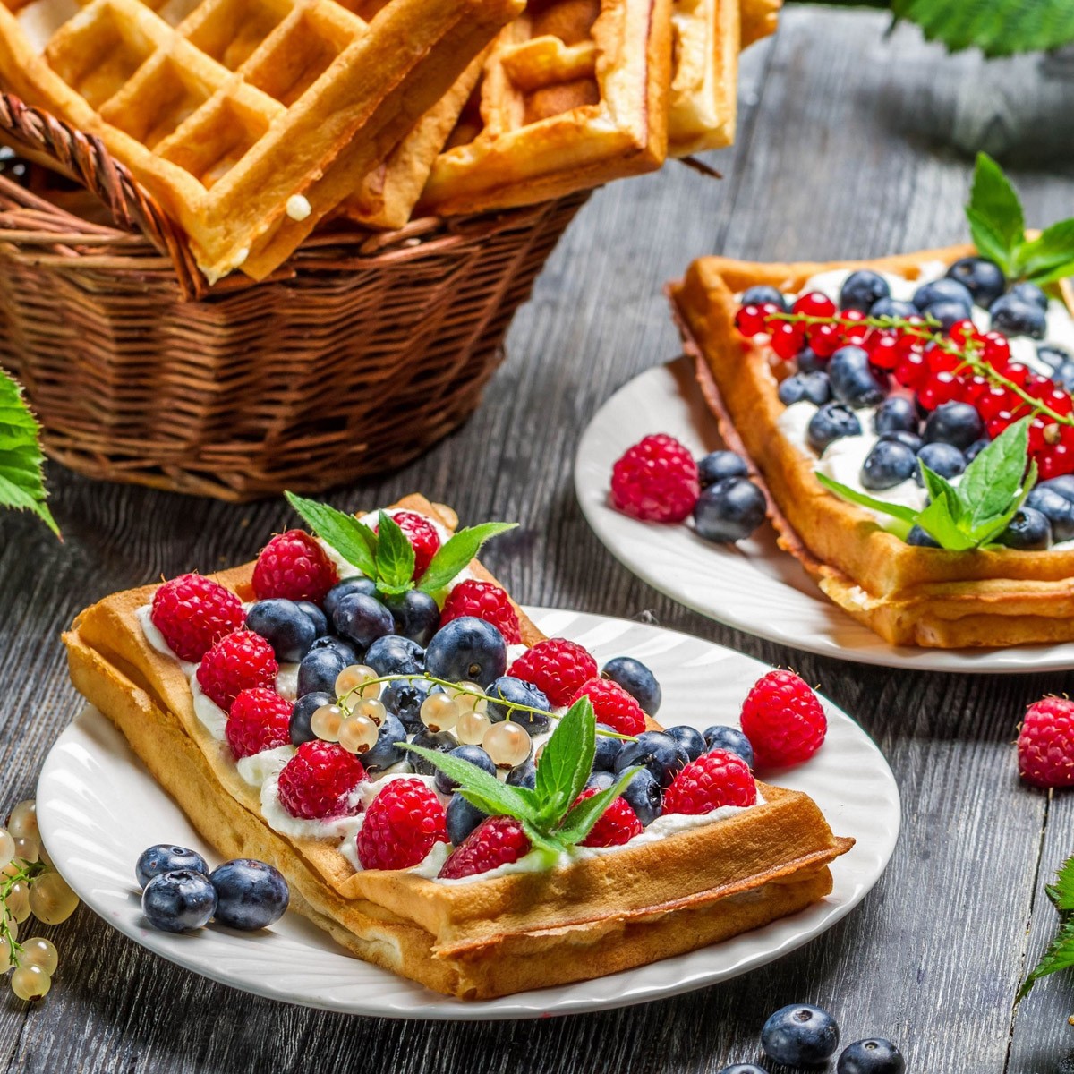 Waffles topped with mixed berries and mint on white plates beside a wicker basket