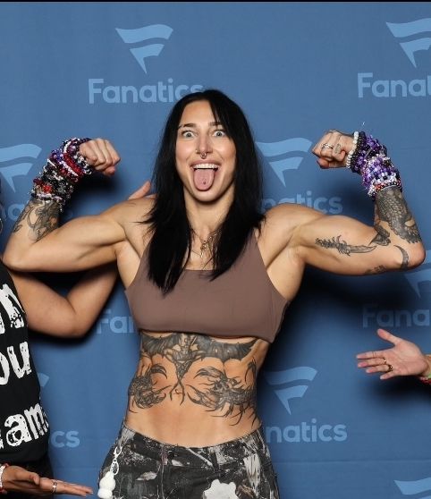 Tattooed woman flexing biceps and making a funny face in front of a Fanatics banner