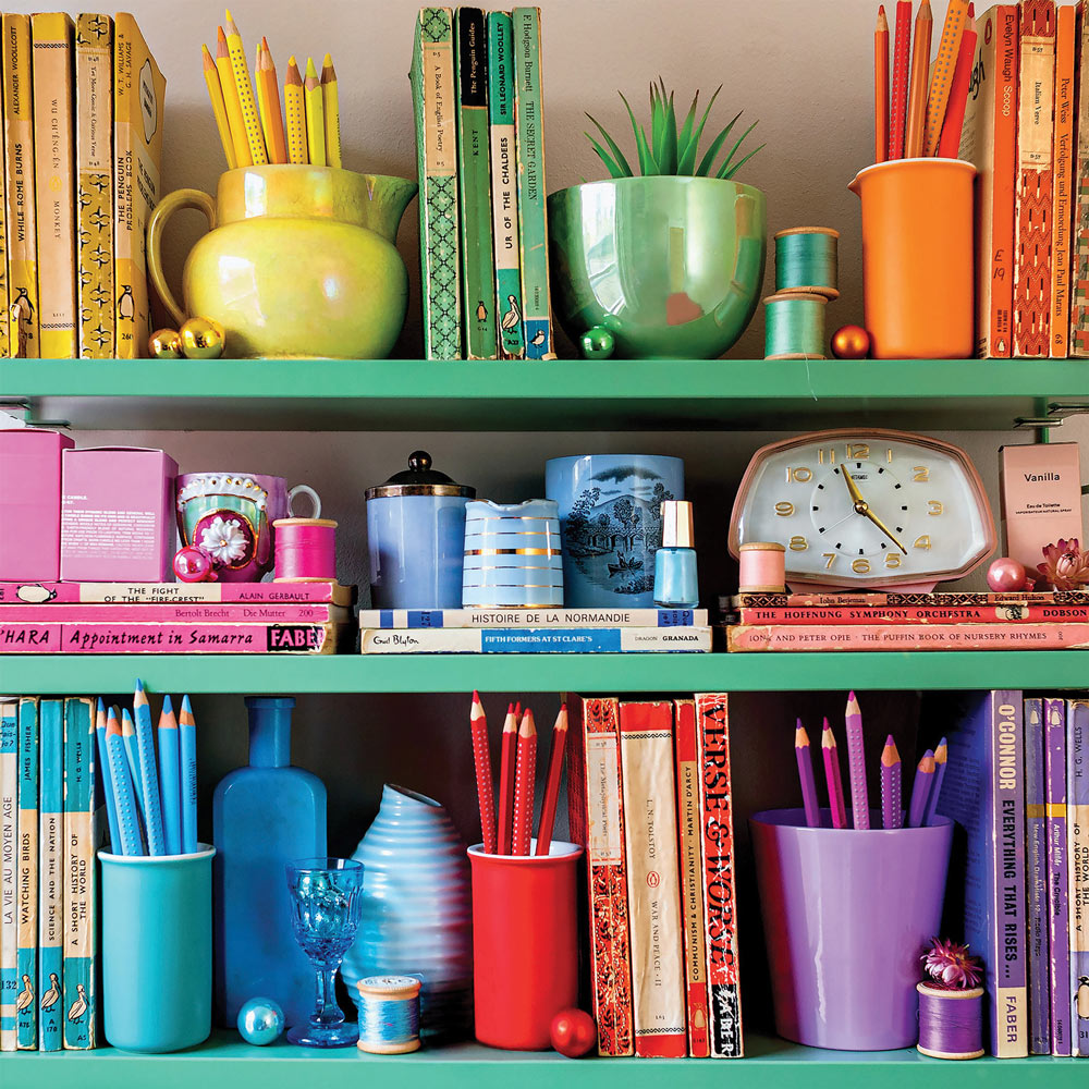 Three shelves with colorful books, cups, clocks, and office supplies organized by color