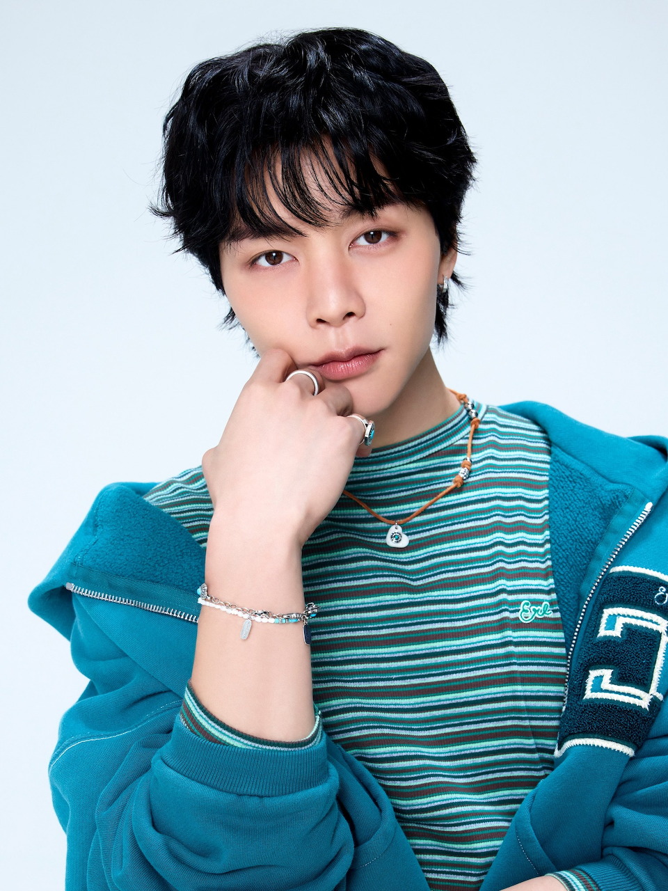 Young East Asian man in teal striped shirt and hoodie, hand near chin, wearing silver bracelet and necklace