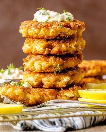 Stack of five golden crab cakes with white sauce and herbs, lemon wedges on silver plate