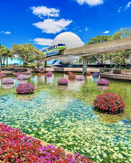 White geodesic dome structure with colorful flower beds in foreground and monorail track, blue sky behind