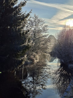 Frost-covered trees along a still creek with mirror-like reflections on a sunny winter day