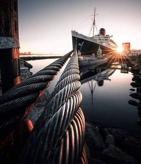 Thick steel mooring cables leading to a large docked ship at sunset with sun starburst and water reflections