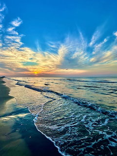 Golden sunrise over ocean with waves washing onto sandy beach beneath blue sky with wispy clouds