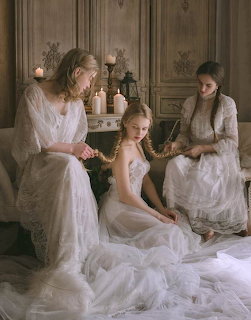 Three women in flowing white lace dresses in an ornate room with candles
