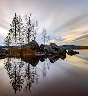 Rocky island with bare trees reflected in calm lake water at sunset with mountains in background