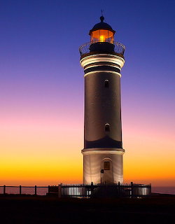 White lighthouse with glowing light at sunset against vibrant purple and golden sky