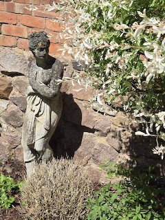 Stone lady statue in a garden beside a brick wall and shrub