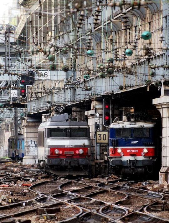Two electric locomotives at a French train station with overhead wires and catenary infrastructure