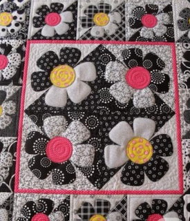 Patchwork quilt block with black and white daisy appliqués and pink accents on a quilted background.