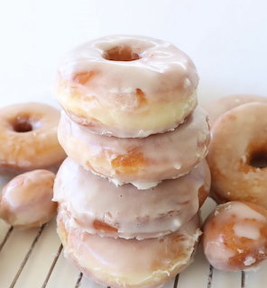 Stack of four glazed donuts with white frosting on wooden slats, more donuts visible at base