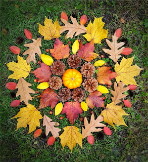 Symmetrical mandala made of fall leaves, acorns, and pumpkin arranged in concentric circles on green grass