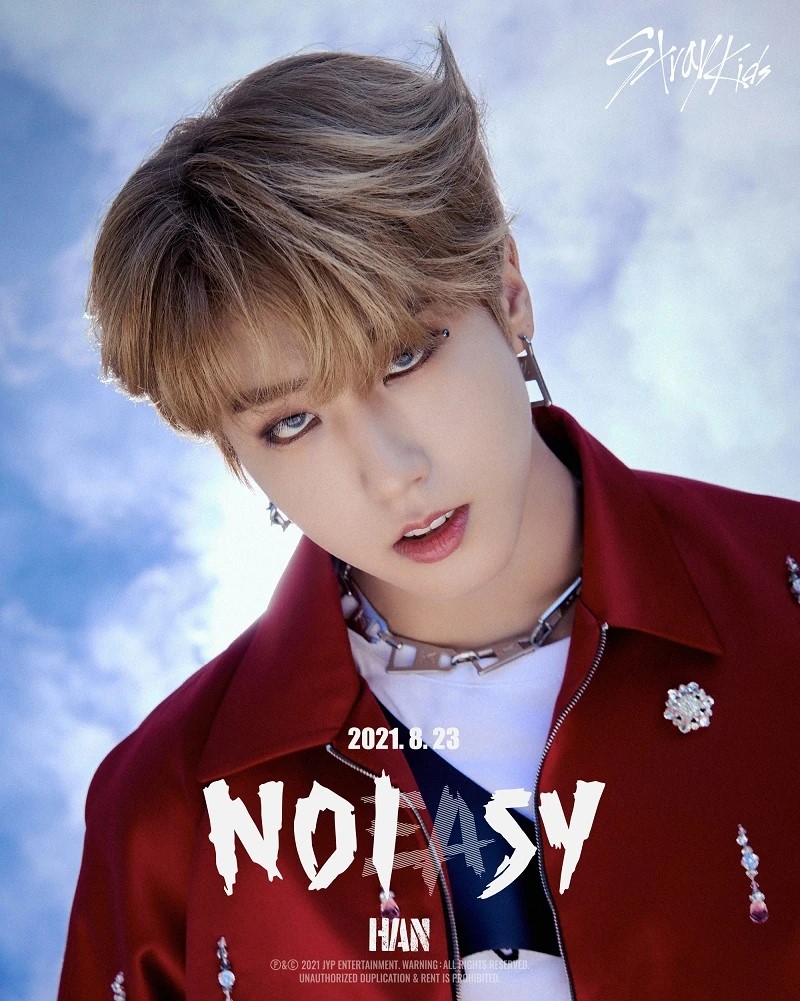 Young man with blond hair wearing a red embellished jacket posing against a cloudy blue sky
