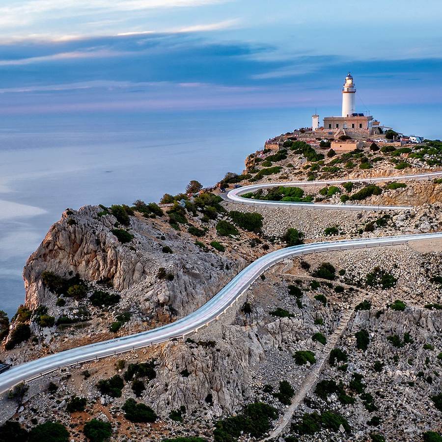 White lighthouse on rocky cliff overlooking ocean with winding coastal road in Majorca Spain