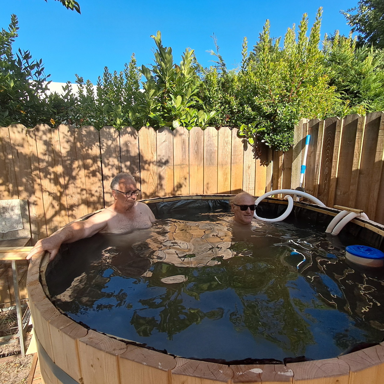 Two adults sitting in a round wooden hot tub in a sunny backyard
