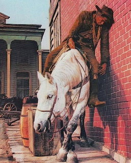 Cowboy in black clothing sitting on white horse in front of red brick building in western film scene