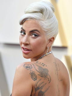 Lady Gaga looking over her shoulder with platinum hair, back tattoos, and a jeweled necklace