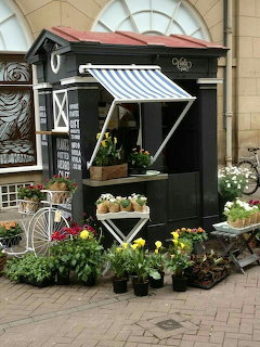 Black wooden flower market stall with striped awning, poted plants, and a white bicycle on cobblestones