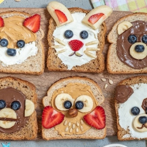 Six slices of toast decorated as cute animal faces with fruit, seeds, and spreads
