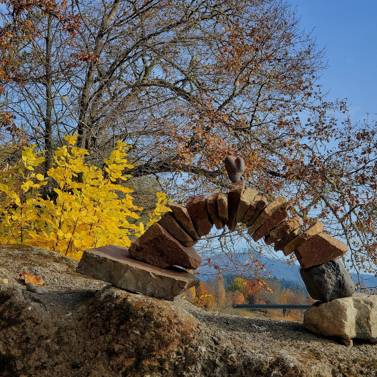 Balanced arch of stacked stones on rocks with autumn trees and blue sky