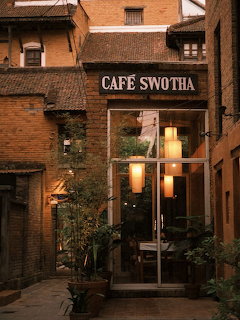 Brick cafe entrance with a glass door, warm lights, and a Cafe Swotha sign in a narrow alley