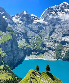 Turquoise alpine lake below rocky snow-dusted mountains and a grassy ridge with two trees