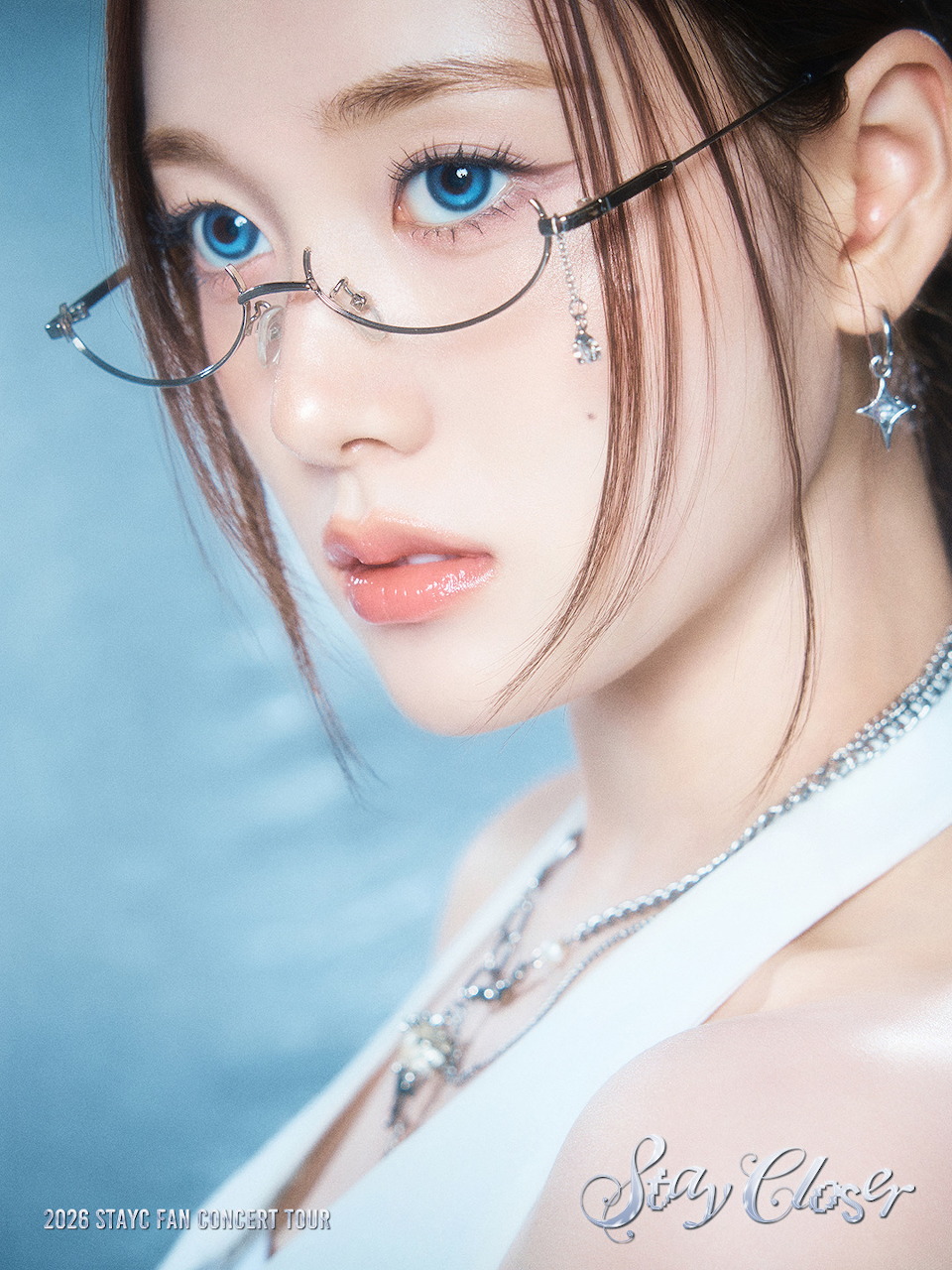 Close-up of STAYC J with blue eyes, rimless glasses, silver jewelry, and a white top.