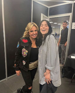 Two smiling women posing together backstage, one in a patched black jacket and the other in a grey shirt