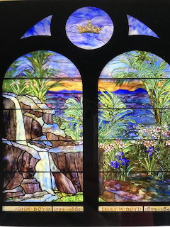 Arched stained glass window showing a waterfall, trees, flowers, and a sunset over water