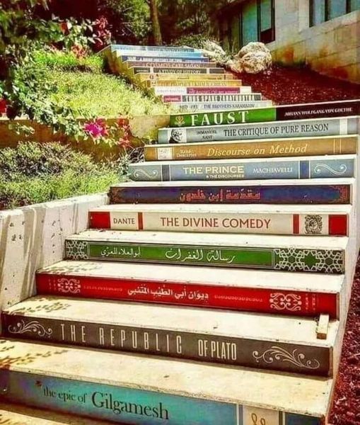 Stone stairs constructed from stacked books ascending between buildings with plants on both sides