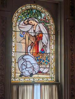 Arched stained glass window depicting an angel in flowing robes with colorful floral border in a castle interior