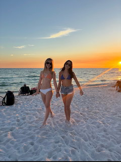 Two women walking together on a white sand beach at sunset with ocean in background