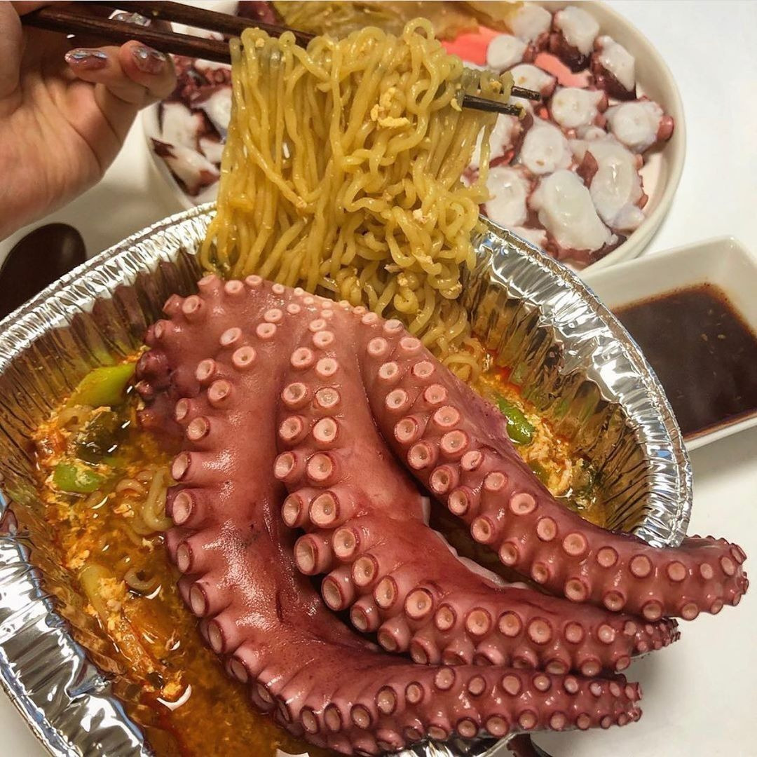 Chopsticks lifting yellow noodles from spicy ramyun topped with pink octopus tentacles