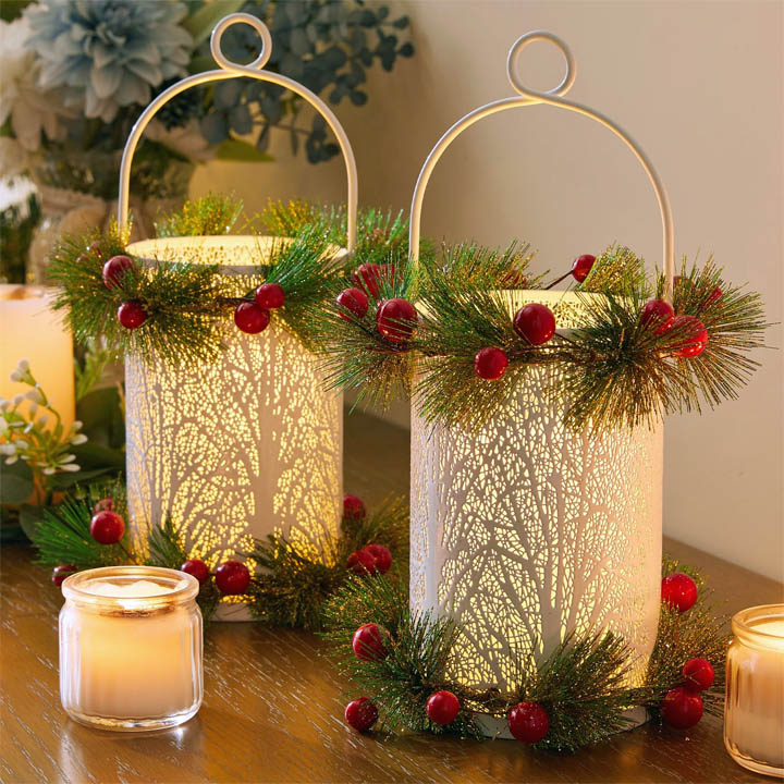 Two illuminated white lanterns decorated with pine branches, red berries, and gold handles on a holiday table