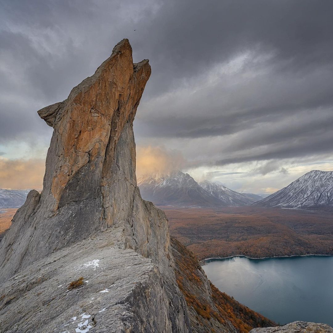 Tall rock spire on a mountain ridge above a lake and snowy peaks under cloudy skies