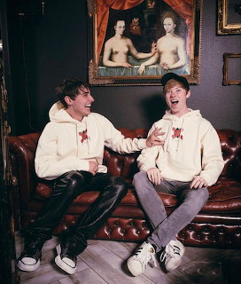 Two men in white hoodies sitting on burgundy couch laughing in room with vintage portrait painting