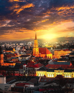 Historic city skyline at sunset with a tall church steeple, red rooftops, and glowing clouds
