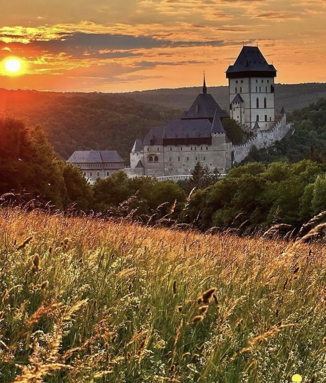 Karlstejn Castle above a grassy meadow at sunset with trees and distant hills
