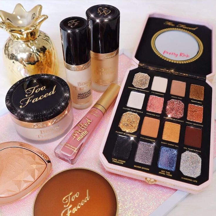 Assorted makeup products on a pink surface, including eyeshadow palette, foundation, powder, and lip gloss