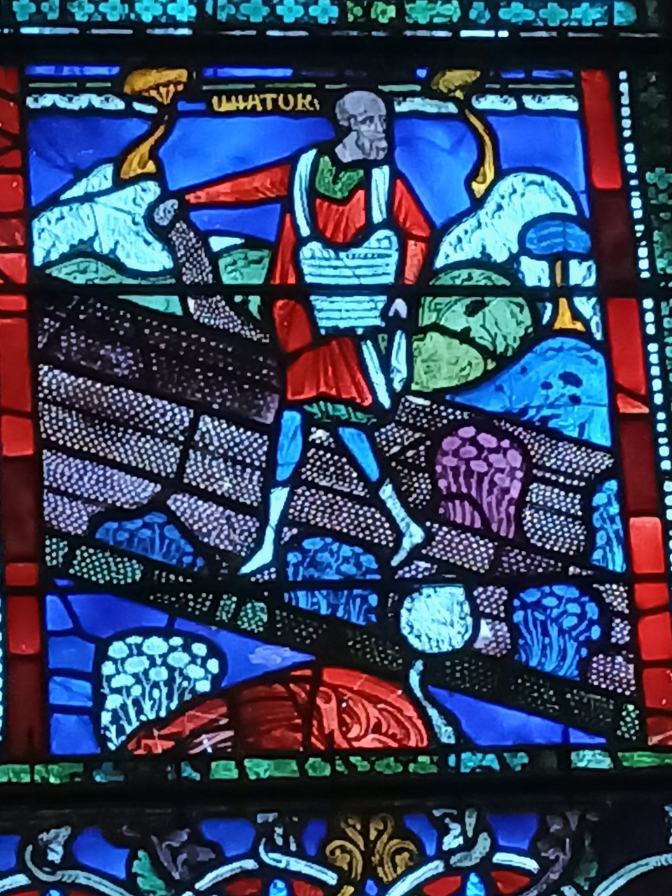 Stained glass window showing a figure in red and blue robes sowing seeds in a field with decorative border