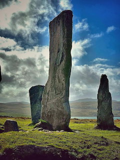 Tall weathered standing stones on a grassy hillside under cloudy blue sky