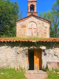 Small stone church with bell tower and wooden door behind a rustic stone wall and trees