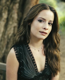 Brunette woman with long wavy hair in a dark v-neck top posing outdoors with greenery behind her