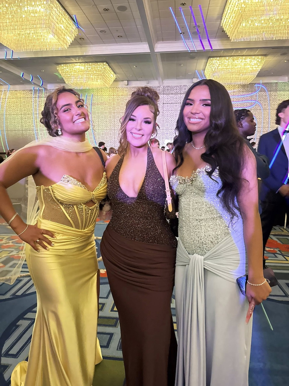 Three young women in gold, brown, and silver prom dresses posing together at a formal event