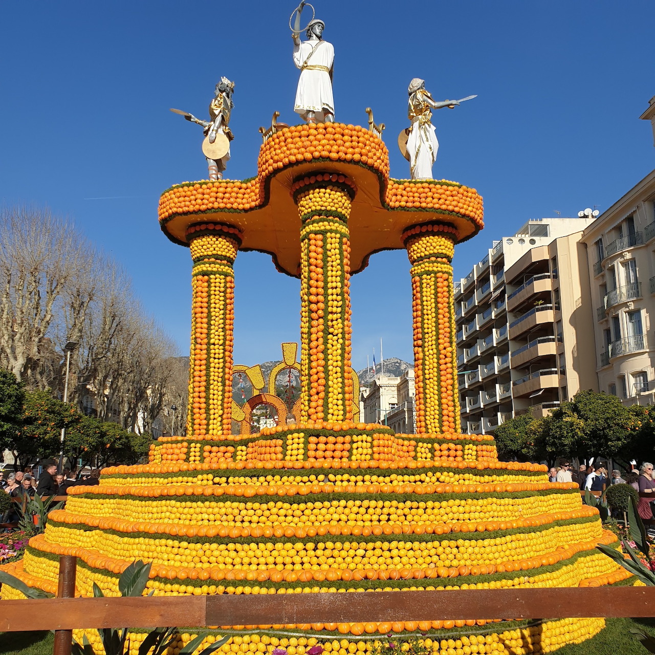 Large decorative structure built from oranges and lemons with columns and white statues on top at outdoor festival