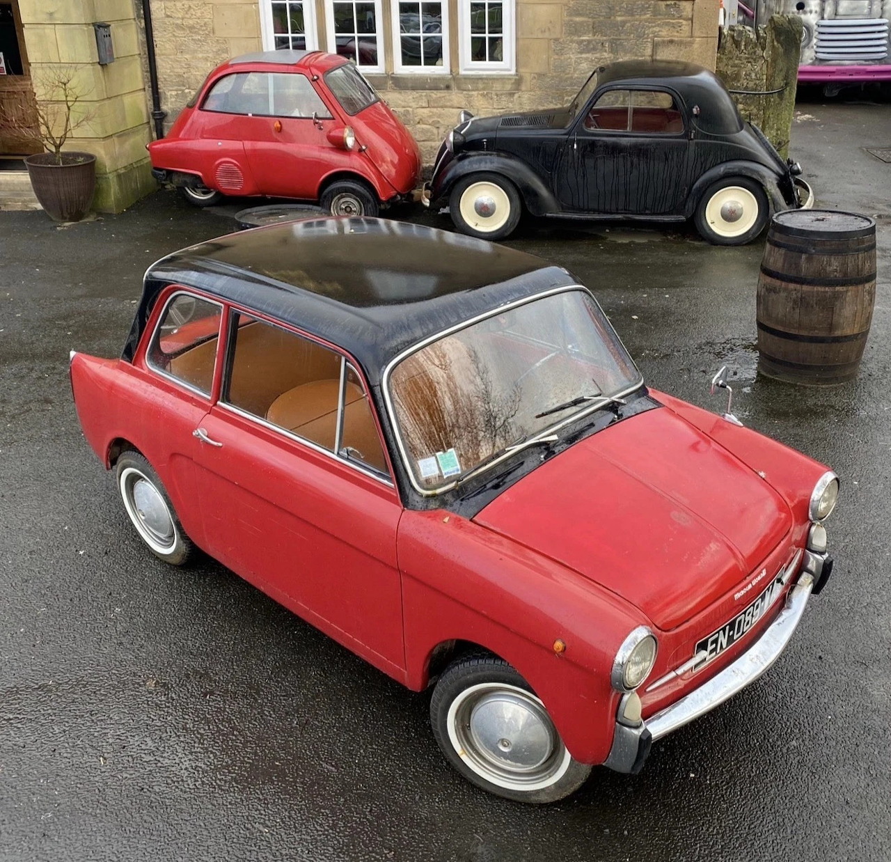 Red vintage Autobianchi Bianchina parked in a courtyard with other classic cars behind it