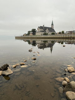 Stone church and steeple reflected in a calm lake with rocks in shallow water under a gray sky