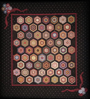 Hexagonal patchwork quilt with brown, pink, and cream fabrics in geometric arrangement on black background with red floral border